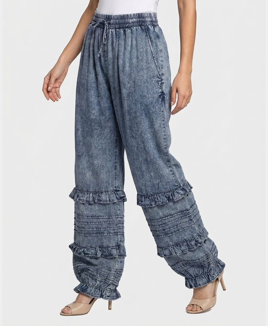 A model wearing wide-leg, high-waisted blue denim jeans with tiered ruffle details on the lower legs, styled with beige high-heeled sandals against a light gray studio background.